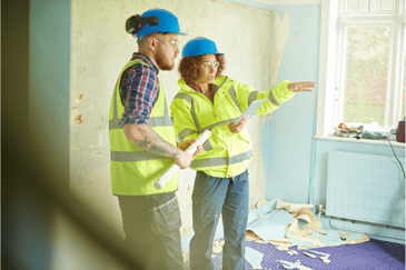 Woman pointing to the side of the room, giving instructions to another builder holding an iPad with construction software on 