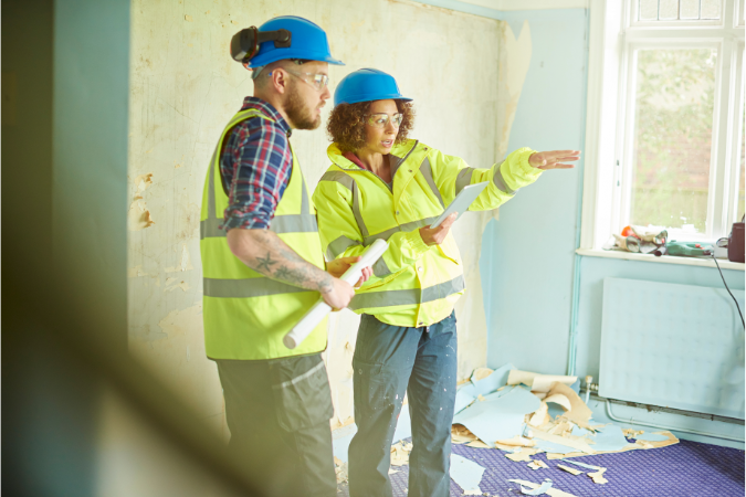 Woman pointing to the side of the room, giving instructions to another builder holding an iPad with construction software on
